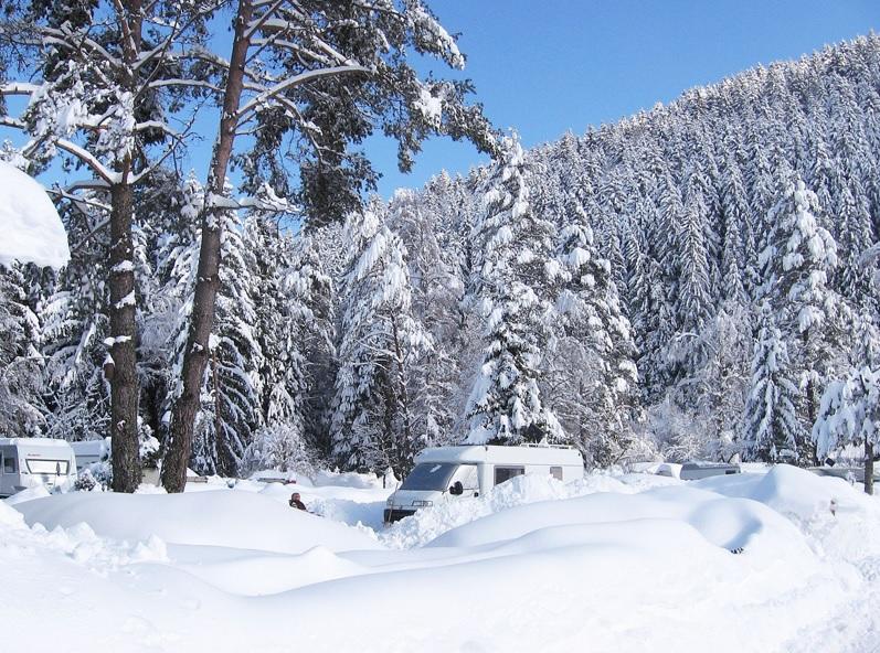 Zwei schneebedeckte Wohnmobile auf verschneitem Boden, umgeben von schneebedeckten Nadelb&auml;umen, bewaldeter Berg im Hintergrund und blauer Himmel