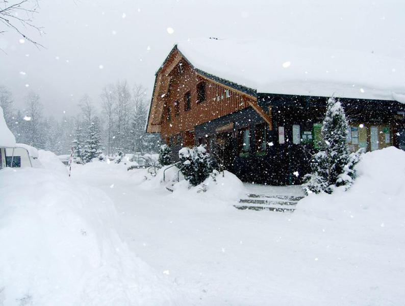 Holzhaus mit verschneitem Dach und Eingangsbereich, fallender Schnee, schneebedeckte B&auml;ume und hohe Schneehaufen im Vordergrund