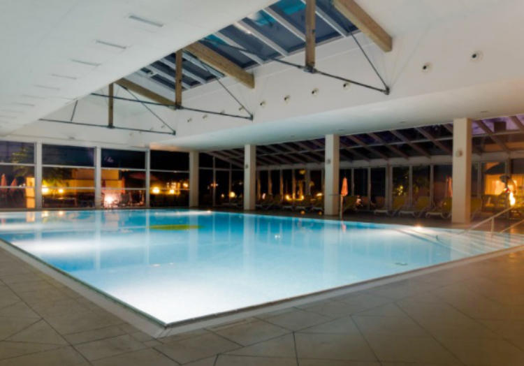 Rechteckiges, beleuchtetes Hallen-Schwimmbecken mit gro&szlig;en Glasfenstern, sichtbaren Deckenbalken und gefliester Umrandung