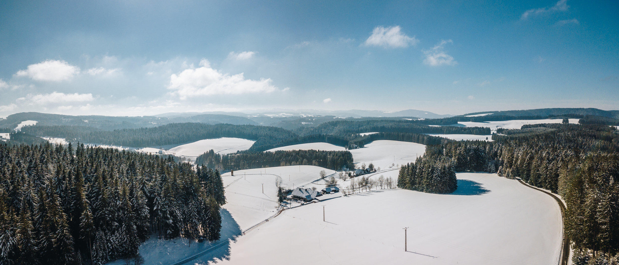 Panoramablick auf schneebedeckte Felder und H&uuml;gel mit Nadelw&auml;ldern und einzelnen Geb&auml;uden unter blauem Himmel