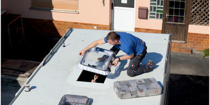 Person kniet auf dem wei&szlig;en Dach eines Wohnmobils und &ouml;ffnet eine Dachluke; Kopf einer zweiten Person schaut durch die Luke; Werkzeugkiste und Materialien auf dem Dach; Hausfassade im Hintergrund