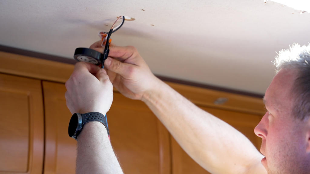 Person mit kurzem Haar und Smartwatch hält freiliegende Drähte und eine runde Deckenbefestigung in einer Öffnung der Decke über Holzschränken