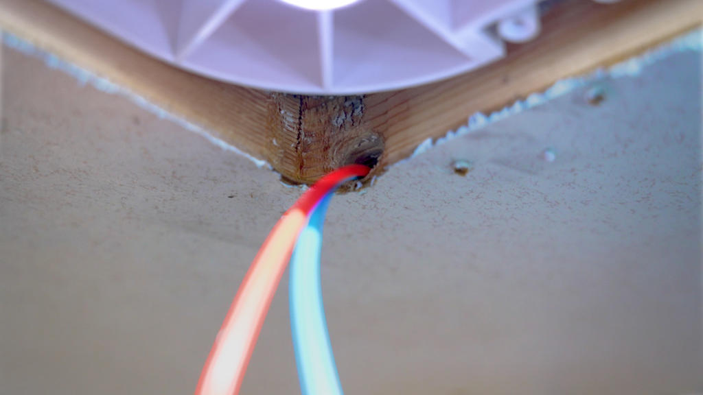 Zwei isolierte Kabel (rot/orange und hellblau/weiß) treten durch ein Loch in einem Holzstück an der Decke neben einer runden weißen Deckenhalterung; Deckenplatten mit sichtbaren Schrauben