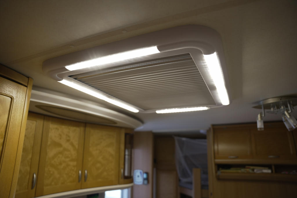 Decke mit rechteckiger Lüftungsabdeckung mit Lamellen, zwei langen Deckenleuchten, Holzschränken mit Griffen, kleinem weißem Wandgerät und abgedecktem Bereich im Hintergrund