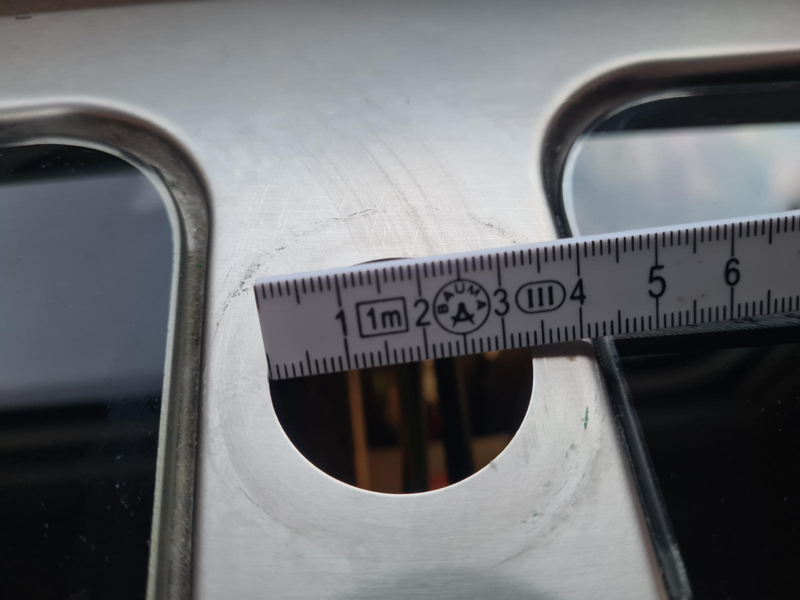 Nahaufnahme einer Metallplatte mit zwei länglichen Aussparungen, einem runden Loch und einem Metalllineal, das über dem Loch liegt; darunter zwei schwarze rechteckige Flächen sichtbar