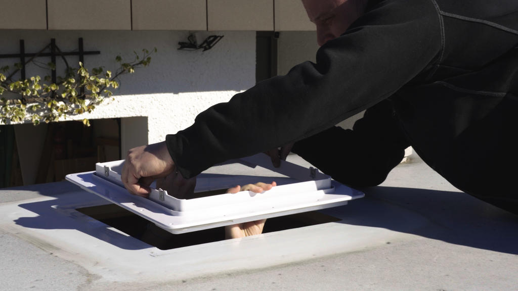 H&auml;nde einer Person in dunklem Oberteil heben einen wei&szlig;en, quadratischen Dachluken-Deckel; im offenen Rahmen sind zwei nackte F&uuml;&szlig;e sichtbar; Dachfl&auml;che und Geb&auml;udefassade im Hintergrund