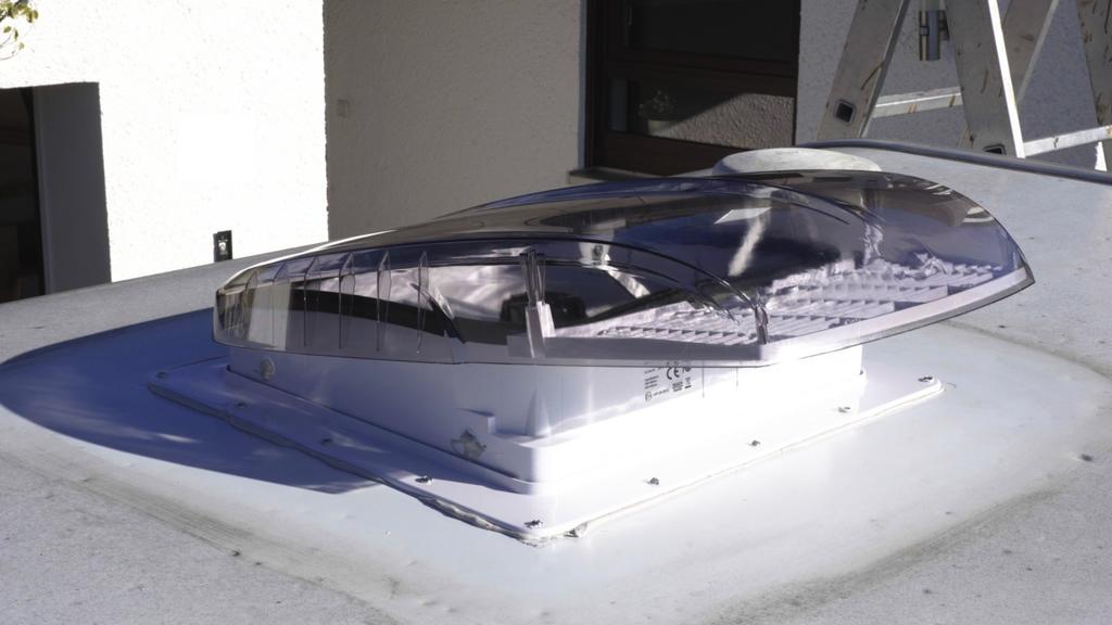 Transparente gewölbte Kunststoff-Dachkuppel auf rechteckiger weißer Basis auf einem Flachdach, im Hintergrund eine weiße Wand mit Fenster