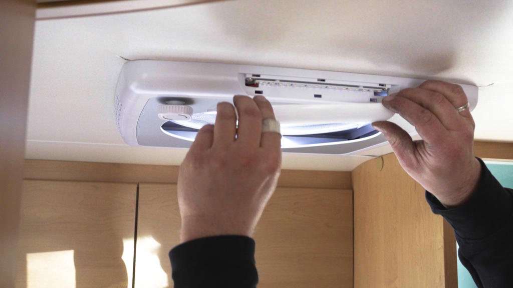 Zwei H&auml;nde halten ein wei&szlig;es, rechteckiges Deckenl&uuml;fter-/Klappenteil an einer hellen Holzdecke; Unterarme mit dunklen &Auml;rmeln sichtbar