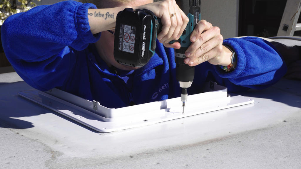 Person in blauem Oberteil h&auml;lt einen Akku-Bohrschrauber &uuml;ber einer wei&szlig;en rechteckigen Dachluke auf einer flachen, grauen Oberfl&auml;che; sichtbare H&auml;nde und ein Tattoo am Unterarm