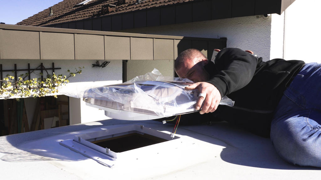 Person legt rechteckige Metallabdeckung &uuml;ber rechteckige &Ouml;ffnung auf Flachdach; tr&auml;gt dunkle Jacke und blaue Jeans; Ziegeldach im Hintergrund