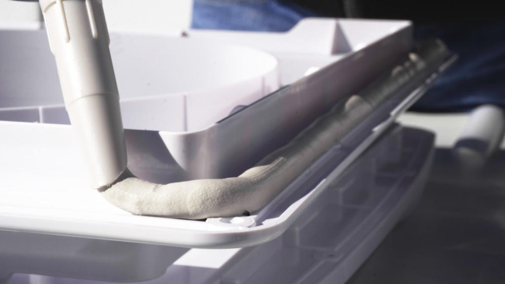 D&uuml;senaufsatz tr&auml;gt graubeigen Dichtstoff entlang der Kante eines wei&szlig;en Kunststoffbeh&auml;lters auf; weitere wei&szlig;e Beh&auml;lter im Hintergrund