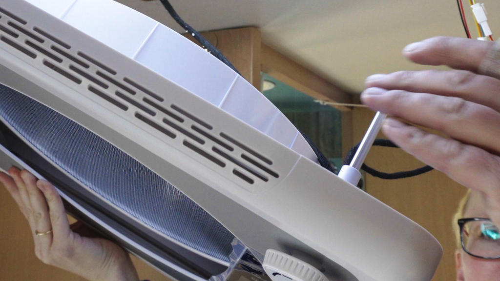 Zwei H&auml;nde halten eine runde wei&szlig;e Leuchte; eine Hand dreht mit einem Schraubendreher an einer Schraube; am rechten Bildrand ist eine Gesichtspartie mit Brille sichtbar; Holzoberfl&auml;che im Hintergrund