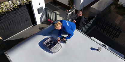 Zwei Personen auf einem flachen wei&szlig;en Dach; eine kniet neben einem offenen Dachfenster, die andere steht in der N&auml;he einer Leiter, im Hintergrund Geb&auml;ude