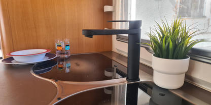 Schwarzer Wasserhahn &uuml;ber schwarzem Glaskeramik-Kochfeld; rechts auf der Fensterbank ein wei&szlig;er Blumentopf mit gr&uuml;ner Pflanze; links gestapelte wei&szlig;e Schale und Teller mit rotem Rand sowie zwei kleine Flaschen; Holzverkleidung und Fensterrahmen sicht