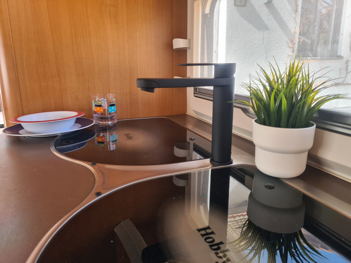 Schwarzer Wasserhahn &uuml;ber schwarzem Glaskeramik-Kochfeld; rechts auf der Fensterbank ein wei&szlig;er Blumentopf mit gr&uuml;ner Pflanze; links gestapelte wei&szlig;e Schale und Teller mit rotem Rand sowie zwei kleine Flaschen; Holzverkleidung und Fensterrahmen sicht