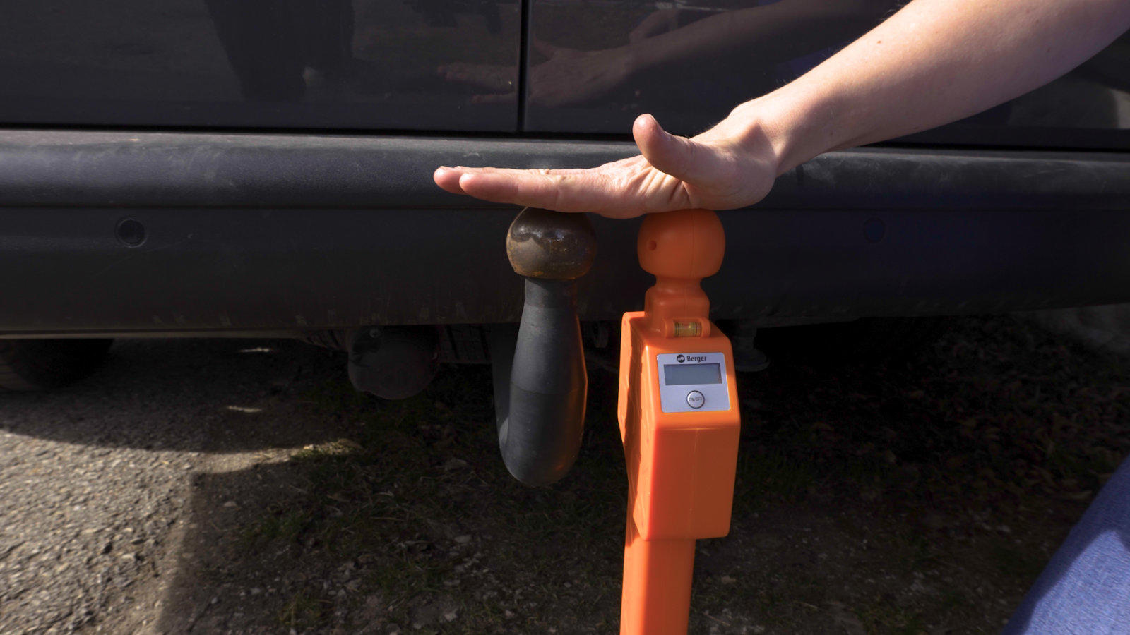 Rechte Hand auf schwarzer Anh&auml;ngerkugel neben orangefarbener Anh&auml;ngerkupplung unter der Hecksto&szlig;stange eines dunklen Autos