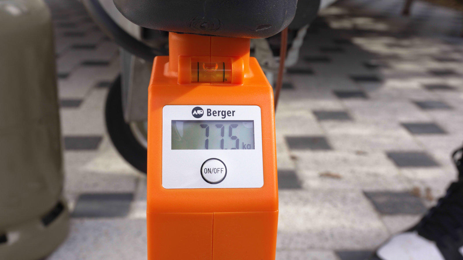 Orangenes Ger&auml;t mit digitalem Display '7.5' und Markenaufdruck 'Berger', auf Pflastersteinen; rechts Teile eines Rads und eines Schuhs sichtbar