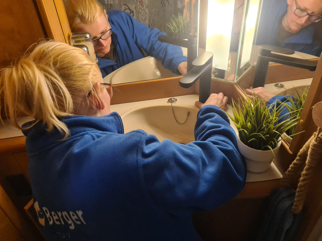 Person in blauer Jacke beugt sich über ein Waschbecken, Hände am Wasserhahn, Blick in den Spiegel, grüne Topfpflanze rechts, Schriftzug Berger auf dem Jackenrücken