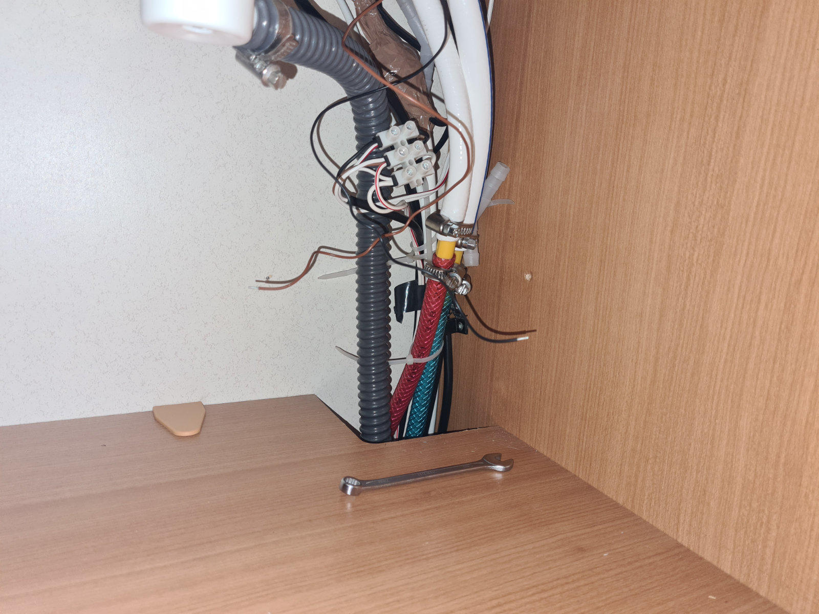 Wei&szlig;e gebogene Oberfl&auml;che oben, schwarze gewellte R&ouml;hre, wei&szlig;e Kunststoffrohre, rote und blaue flexible Schl&auml;uche, mehrere lose Dr&auml;hte mit Klemmenblock, Innenraum eines Holzschranks mit Metallgriff