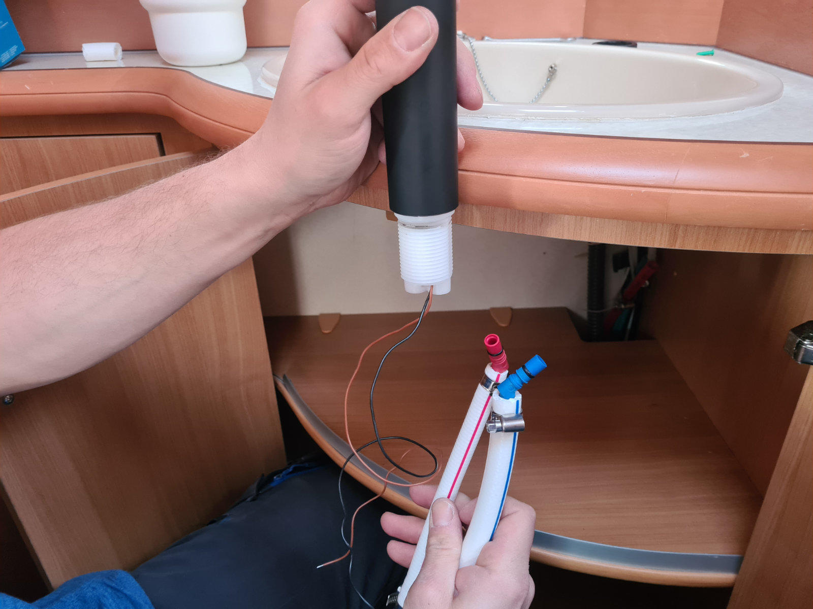Zwei H&auml;nde halten unter einem Waschbecken einen schwarzen zylindrischen Gegenstand mit wei&szlig;er D&uuml;se und ein wei&szlig;es Metallrohr mit roten und blauen Ventilen; offener Holz-Unterschrank, rundes Waschbecken und Armatur sichtbar