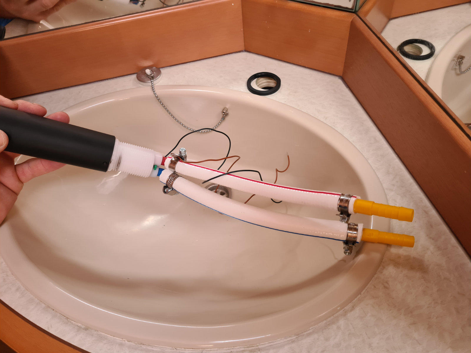 Hand h&auml;lt ein wei&szlig;es Kunststoffrohr mit Kabeln und zwei gelben Schl&auml;uchen &uuml;ber einem wei&szlig;en Waschbecken; brauner Waschtischrand und Spiegel im Hintergrund