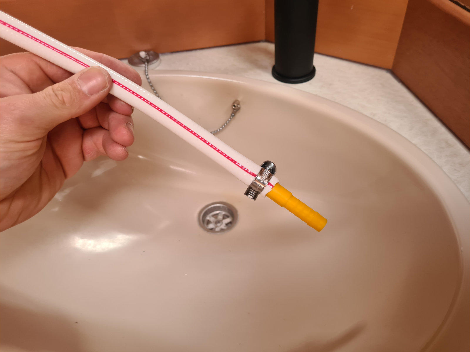 Hand h&auml;lt einen wei&szlig;en Schlauch mit roter L&auml;ngslinie und gelbem Endst&uuml;ck &uuml;ber ein beigefarbenes Waschbecken mit Ablauf und schwarzem Wasserhahnrohr