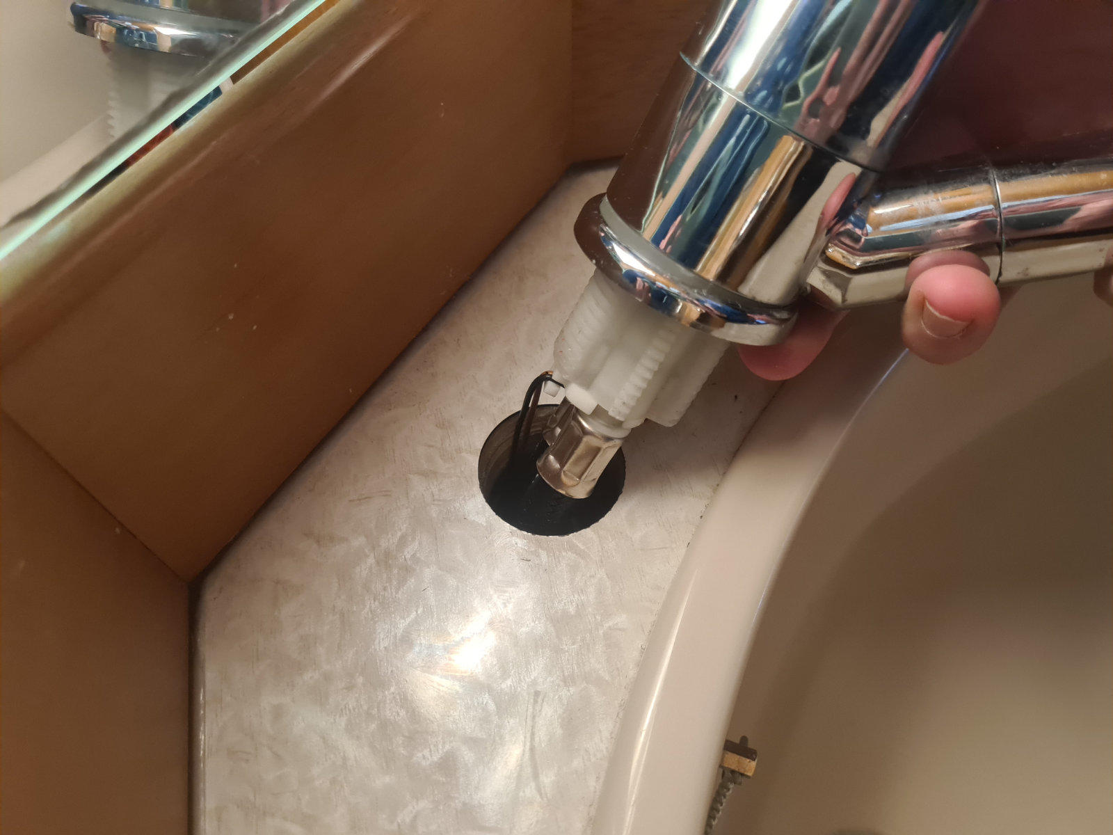 Hand h&auml;lt verchromtes Wasserhahnrohr &uuml;ber Waschbeckenschale mit schwarzem Abluss und beigem Rand; Spiegelkante sichtbar