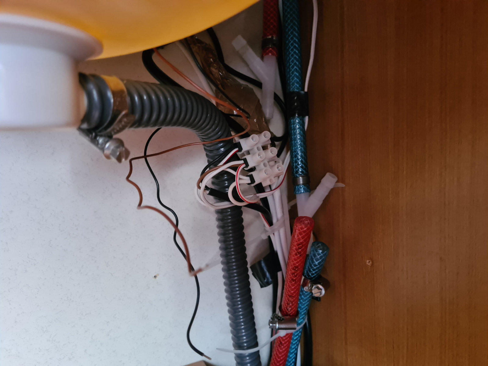 Innenraum eines Unterschranks mit gelber, gew&ouml;lbter Unterseite einer Sp&uuml;le; grauer, flexibler, gerippter Abflussschlauch; wei&szlig;e Kunststoffverbindung; rote und blaue, textilummantelte Schl&auml;uche; schwarze und braune Dr&auml;hte; Kabelbinder; rechte Holzwand