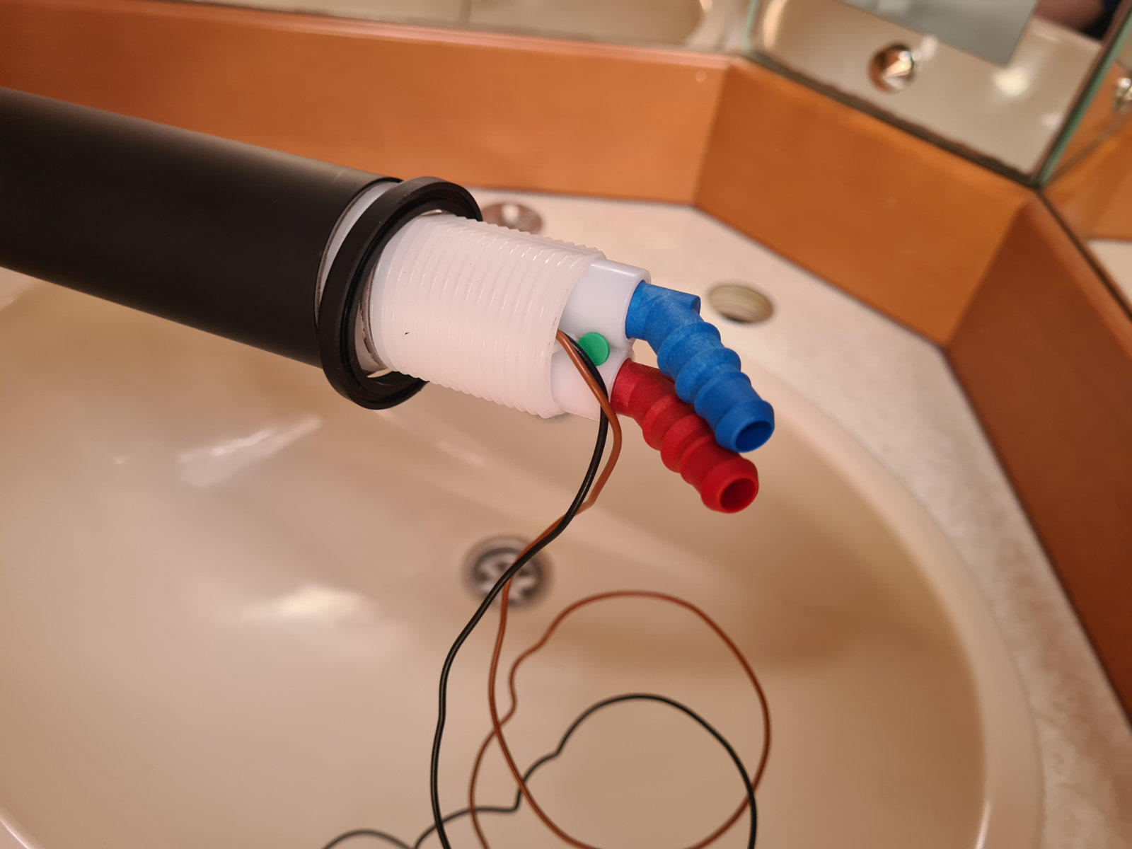 Schwarzes Rohr mit wei&szlig;em Kunststoffanschluss und roten, blauen und gr&uuml;nen Kappen; d&uuml;nne schwarze und braune Dr&auml;hte liegen in einem wei&szlig;en Waschbecken, Holzverkleidung und Metallarmatur im Hintergrund