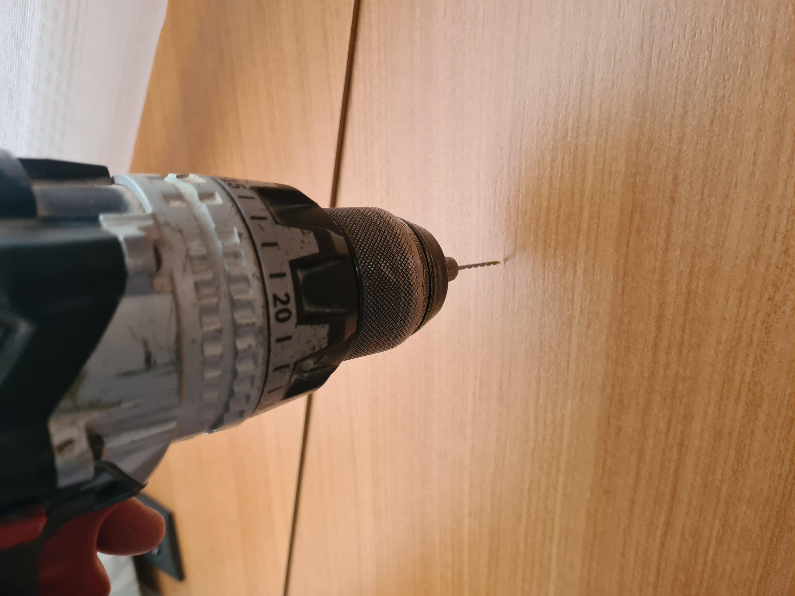 Bohrmaschine mit kleinem Bohrer an einer hellbraunen Holzplatte; links ein heller Vorhangrand sichtbar
