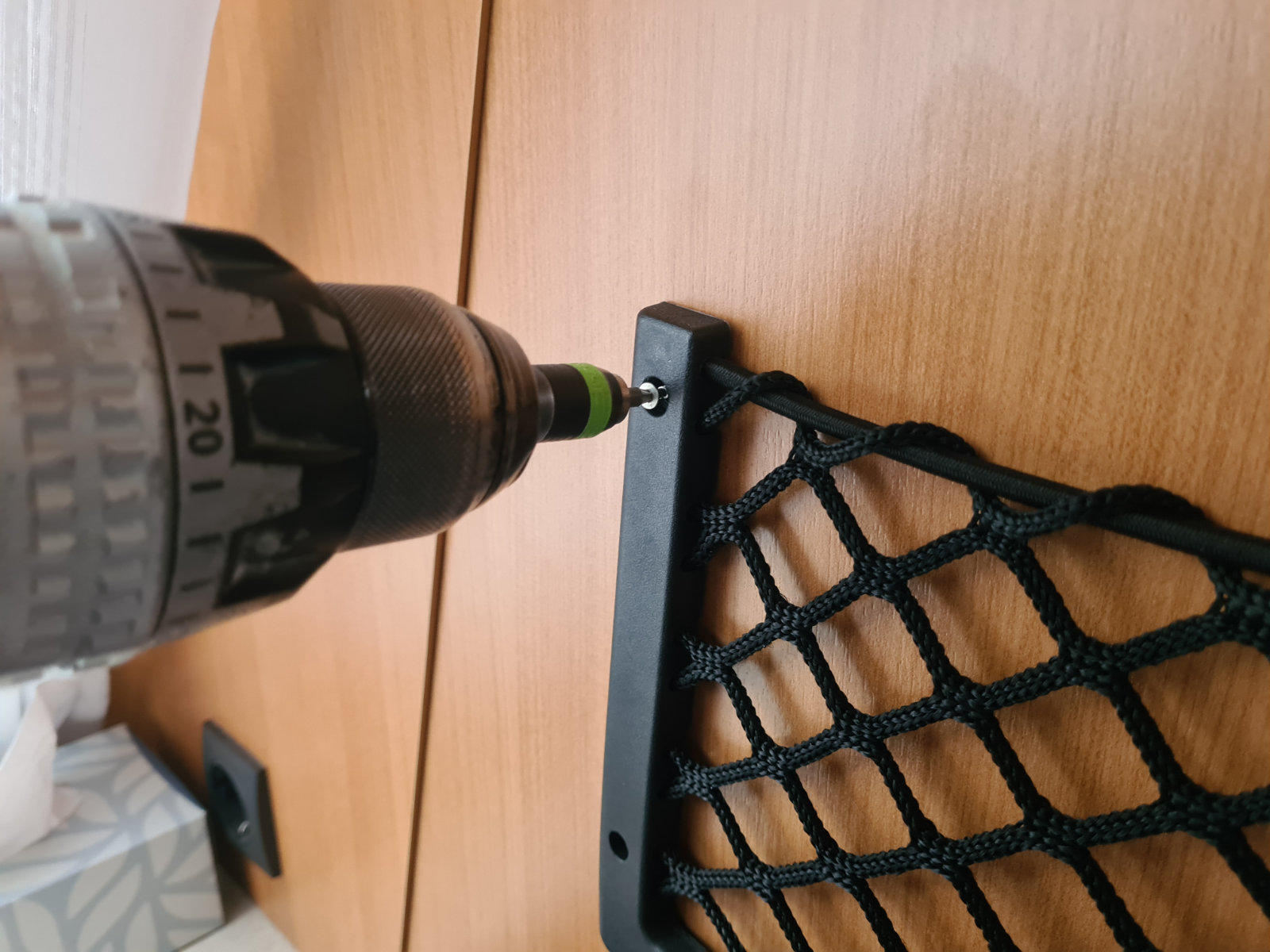 Akkuschrauber mit grünem Bit in einer Schraube an einer Holzschranktür, daneben ein schwarzes elastisches Netz an einem schwarzen Halter