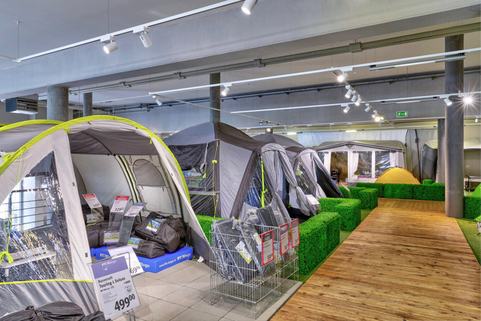 Mehrere aufgestellte Campingzelte auf einer Verkaufsfl&auml;che neben k&uuml;nstlichem Gras und einem Holzsteg unter Deckenbeleuchtung