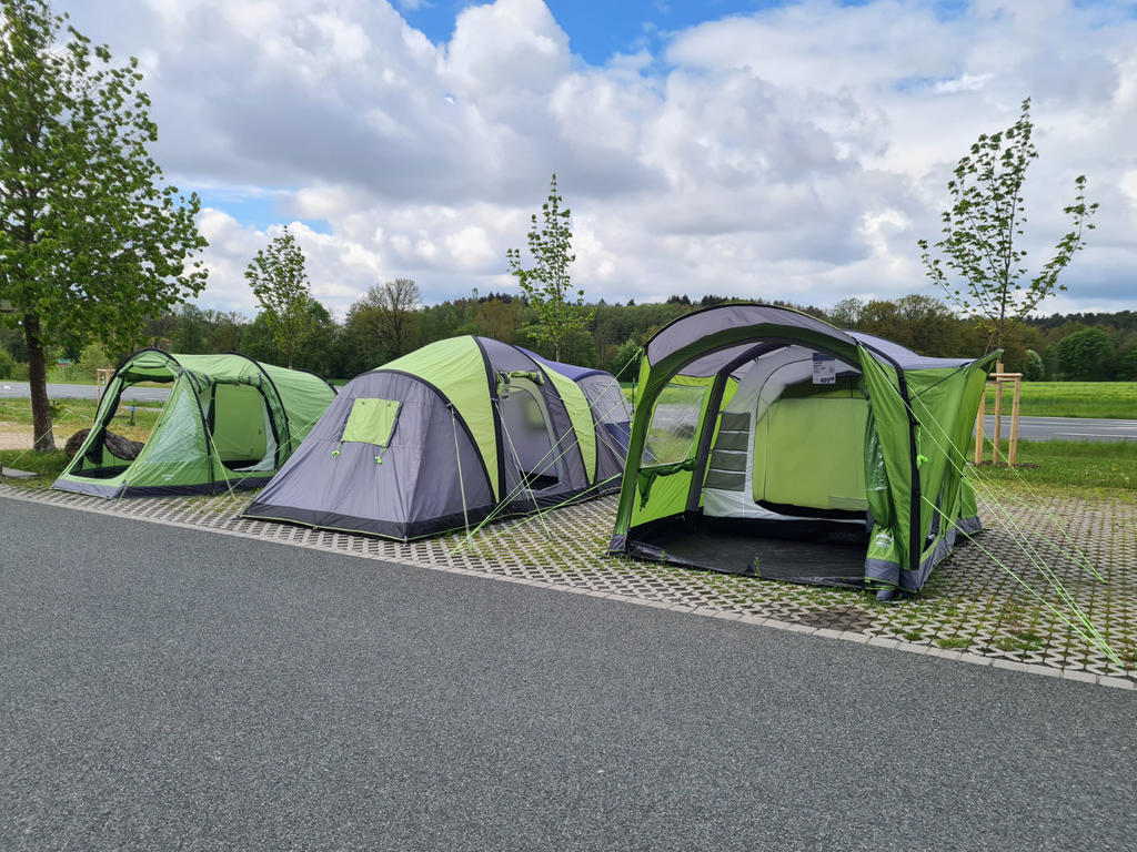 Drei gr&uuml;n-graue Zelte auf einer gepflasterten Stellfl&auml;che neben einer Stra&szlig;e, dahinter B&auml;ume, Rasen und bew&ouml;lkter Himmel