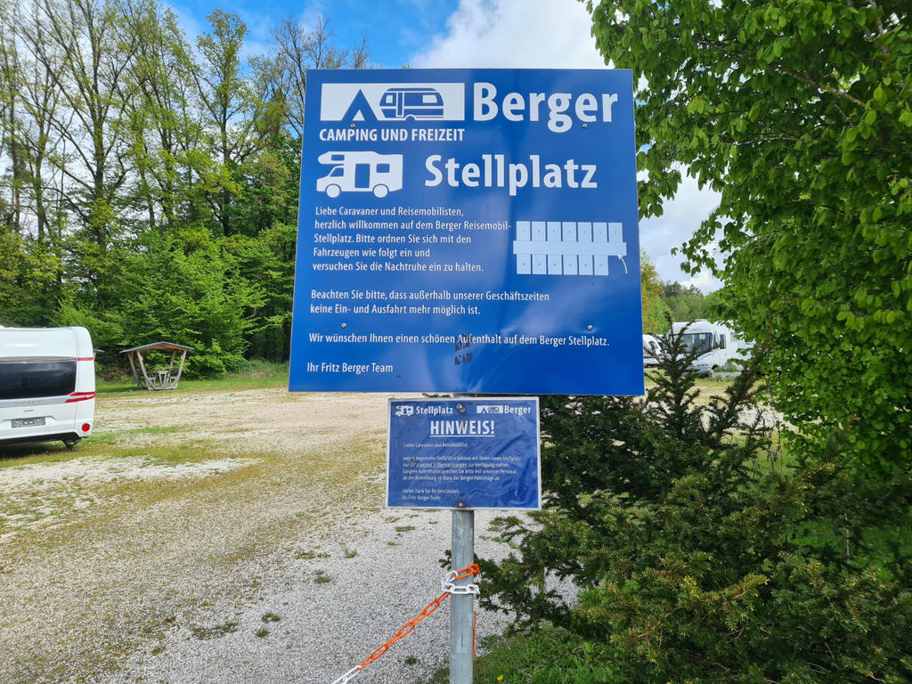 Blaues Schild mit wei&szlig;er Aufschrift Berger Stellplatz und Piktogrammen von Wohnmobilen; kleineres Schild an einem Pfosten darunter, Kiesfl&auml;che, Wohnmobil links, B&auml;ume und Wasserfl&auml;che im Hintergrund, Kette im Vordergrund