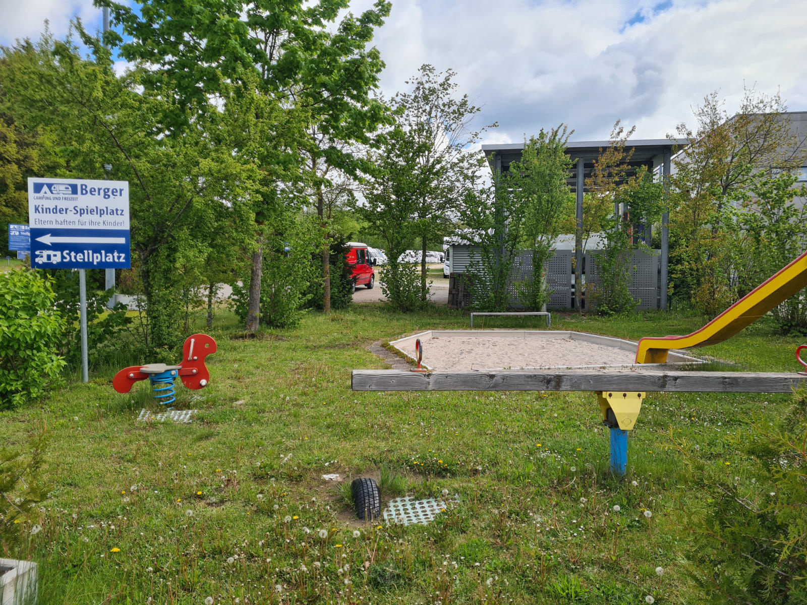 Kleiner Spielplatz mit Sandkasten in Holzumrandung, gelber Rutsche rechts, zwei rote Federwippen links, Grasfl&auml;che mit Entw&auml;sserungsrost im Vordergrund, Hinweisschilder links, B&auml;ume und Geb&auml;ude im Hintergrund, teilweise sichtbare rote Lkw-Kabine