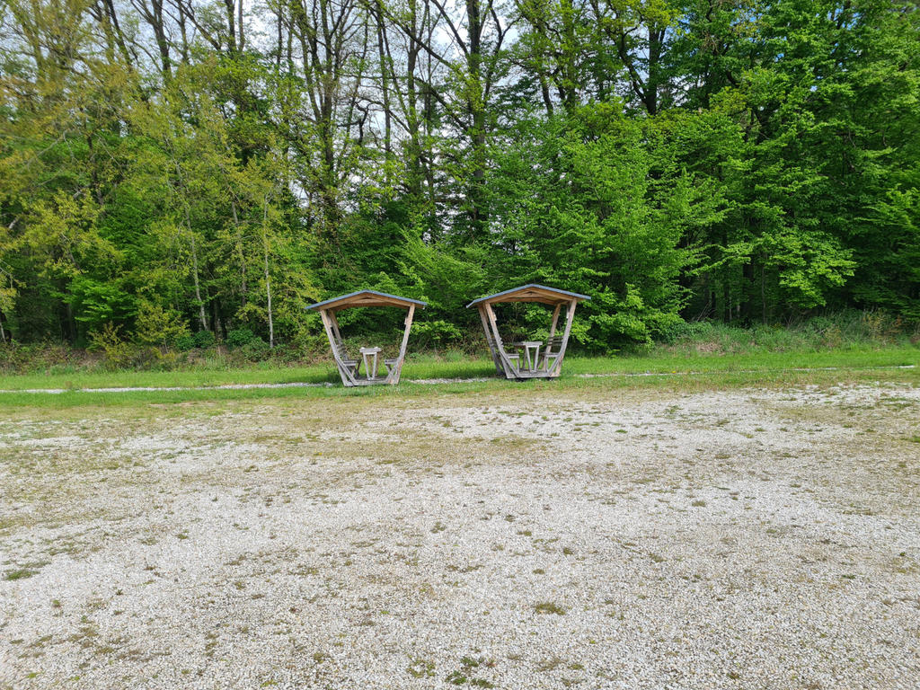 Zwei &uuml;berdachte Holztische mit B&auml;nken nebeneinander vor Baumbestand; Kies im Vordergrund