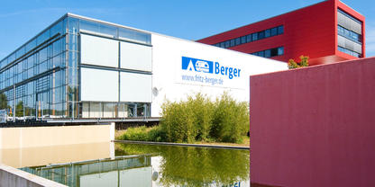 Wei&szlig;es Geb&auml;ude mit Glasfassade und blauem Logo und Schriftzug, rotes Geb&auml;ude im Hintergrund, pinke Wand im Vordergrund, Wasserbecken mit Spiegelung und Bambuspflanzen
