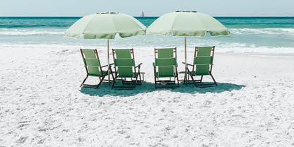 Vier gr&uuml;ne Strandst&uuml;hle unter zwei hellgr&uuml;nen Sonnenschirmen auf wei&szlig;em Sand, t&uuml;rkisfarbenes Meer im Hintergrund