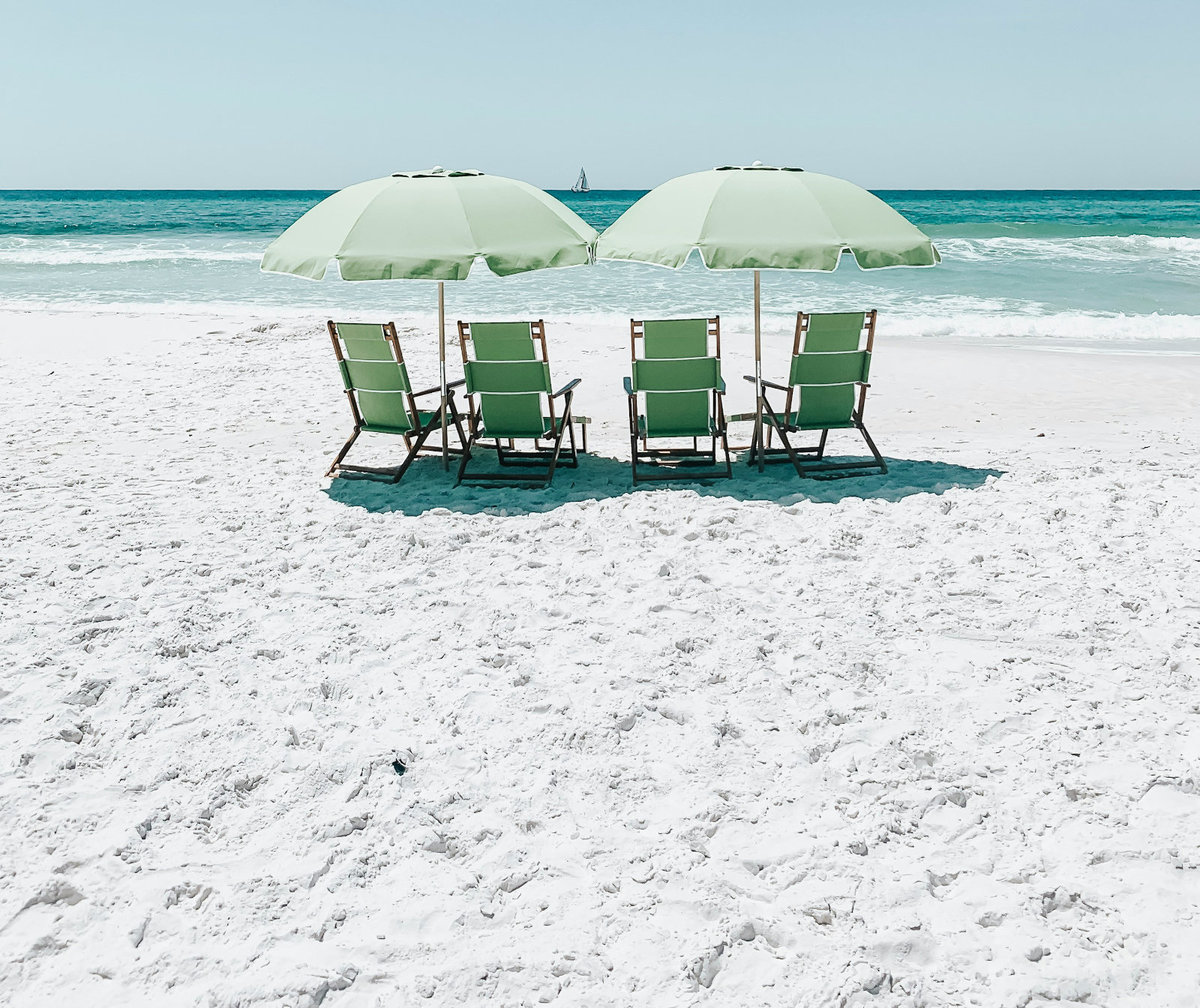 Vier gr&uuml;ne Strandst&uuml;hle unter zwei hellgr&uuml;nen Sonnenschirmen auf wei&szlig;em Sand, t&uuml;rkisfarbenes Meer im Hintergrund