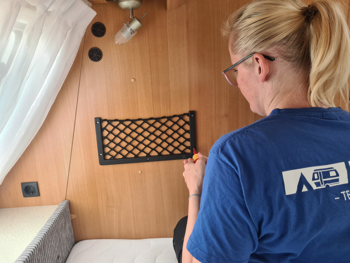Person von hinten mit blondem Pferdeschwanz und Brille, blaues T‑Shirt mit wei&szlig;em Logo, h&auml;lt ein Werkzeug an einer holzvert&auml;felten Wand mit schwarzer Netzablage, Vorhang links, Matratze unten