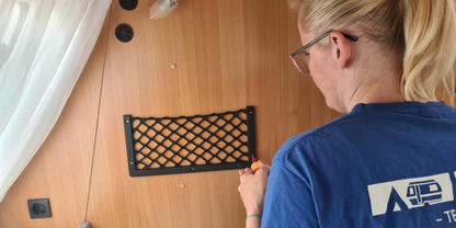 Person von hinten mit blondem Pferdeschwanz und Brille, blaues T‑Shirt mit wei&szlig;em Logo, h&auml;lt ein Werkzeug an einer holzvert&auml;felten Wand mit schwarzer Netzablage, Vorhang links, Matratze unten