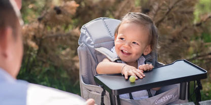 Kleinkind sitzt in einem Hochstuhl im Freien, l&auml;chelt, Hand auf dem Tablett mit einer gr&uuml;nen Traube; unscharfe Person im Vordergrund