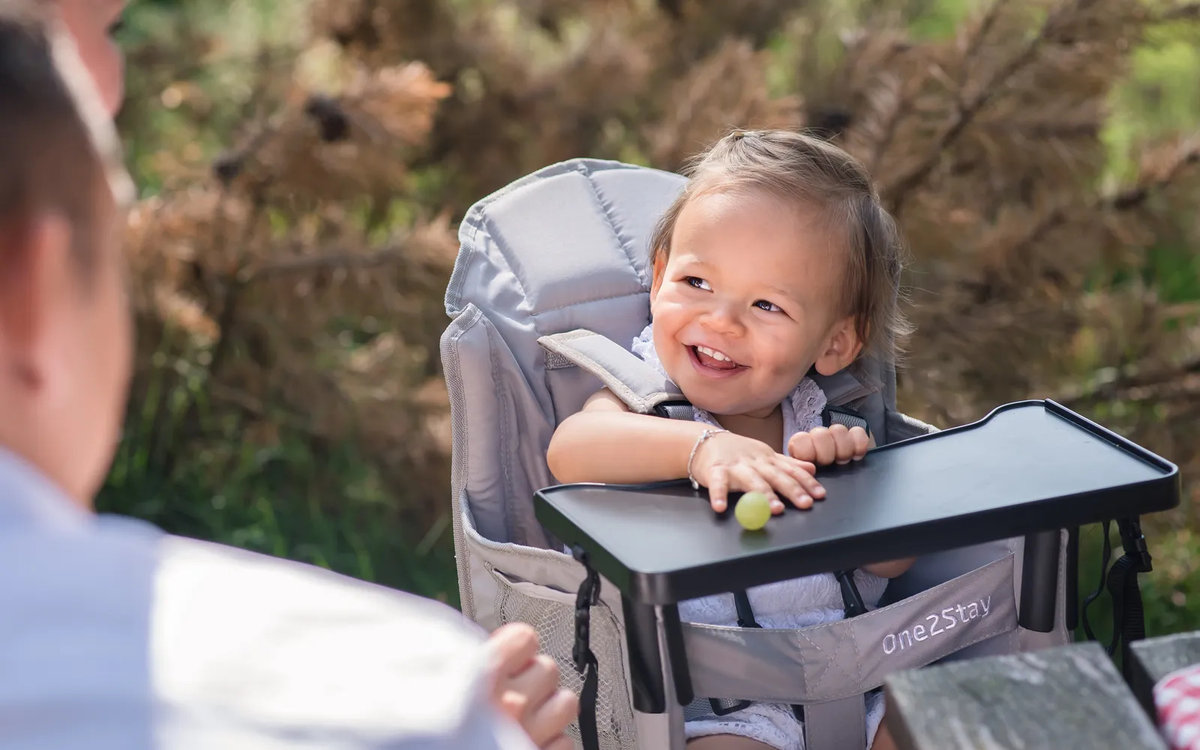 Kleinkind sitzt in einem Hochstuhl im Freien, l&auml;chelt, Hand auf dem Tablett mit einer gr&uuml;nen Traube; unscharfe Person im Vordergrund