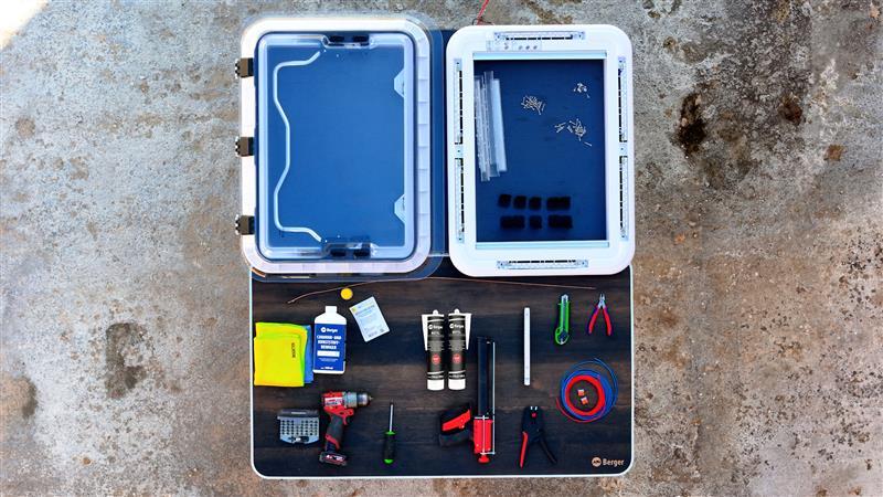 Offener Kunststoffkoffer mit zwei Einsätzen im Deckel; im Kofferboden auf schwarzer Einlage mehrere Werkzeuge und Zubehör: zwei Zangen (grün, rot), zwei Schraubendreher, zwei kleine Metallzylinder, rote Kabelrolle, Flasche mit blauem Etikett, gelbes