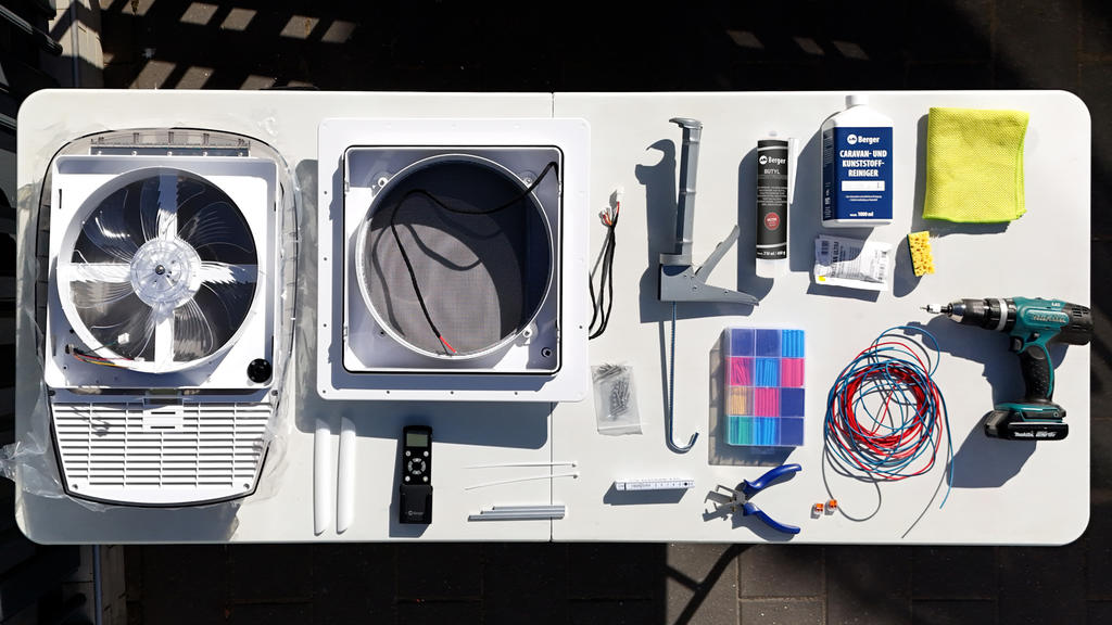 Wei&szlig;er Tisch mit zwei wei&szlig;en Geh&auml;usen (eines mit sichtbarem Propeller), rundem Ventilator, Akku-Bohrschrauber, Kabeln, Schrauben, Zangen, Fernbedienung, Farbk&auml;rtchen, Tube, Flasche und gelbem Tuch