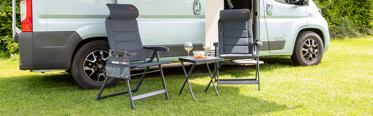 Wei&szlig;es Fahrzeug auf Gras, davor zwei graue Klappst&uuml;hle und ein Klapptisch mit zwei Tassen und weiteren Gegenst&auml;nden