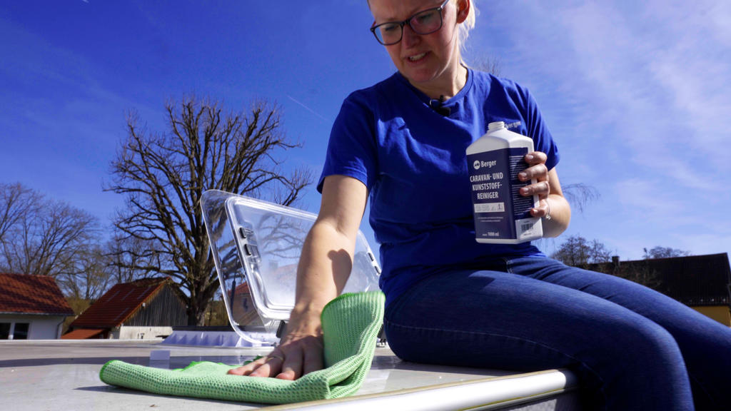 Person in blauem T‑Shirt sitzt drau&szlig;en, wischt eine helle Fl&auml;che mit gr&uuml;nem Tuch und h&auml;lt eine Kartonpackung; im Hintergrund B&auml;ume, H&auml;user und blauer Himmel