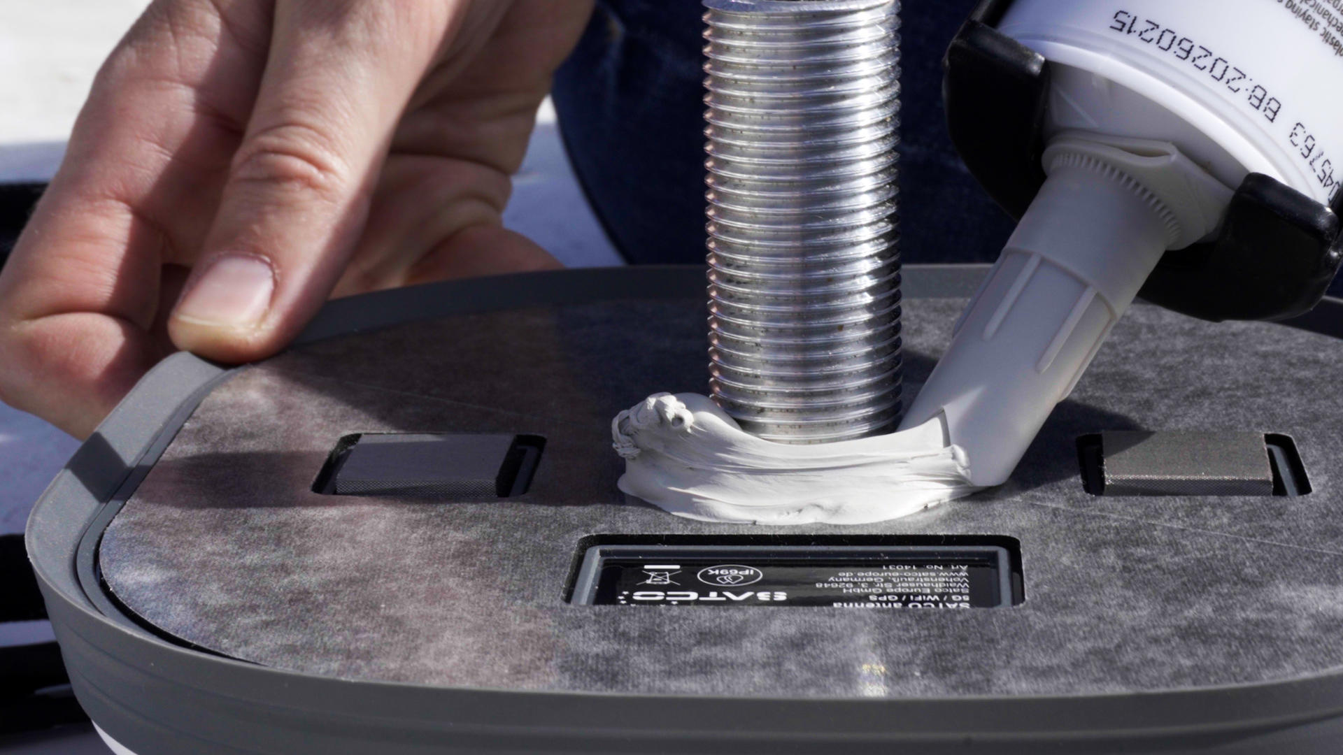 Eine Kartuschenpistole gibt wei&szlig;e Dichtmasse um einen Metallgewindebolzen auf einer grauen Kunststoffbasis ab; eine Hand h&auml;lt die Basis