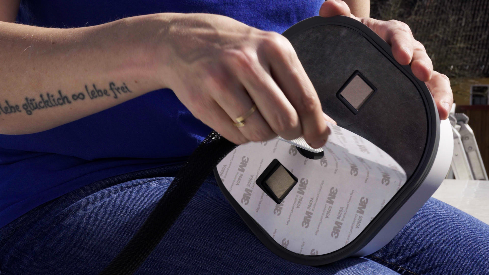 H&auml;nde &uuml;ber einem offenen, ovalen Ger&auml;t mit zwei quadratischen &Ouml;ffnungen; eine Hand h&auml;lt ein herausgenommenes quadratisches Teil. Sichtbar: blaues Oberteil, Jeans, T&auml;towierung am Unterarm, Ring am Finger