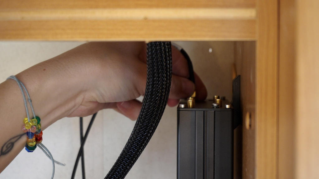 Hand mit buntem Armband und kleinem Tattoo h&auml;lt einen schwarzen geflochtenen Kabelschlauch neben einer schwarzen Ger&auml;tebox mit goldenen Steckern in einem Holzschrank